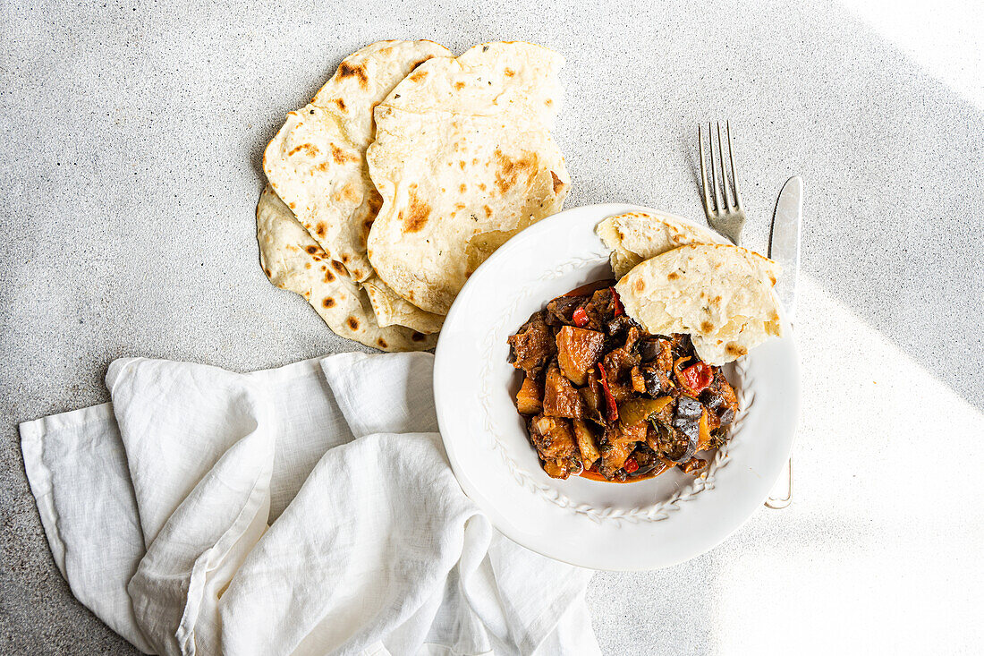 Ein köstlicher hausgemachter Gemüseeintopf mit Auberginen, Tomaten, Kartoffeln, Paprika, Zwiebeln, Knoblauch und Basilikum, begleitet von frisch gebackenem Fladenbrot, dargestellt auf einer weißen Fläche mit einer Leinenserviette