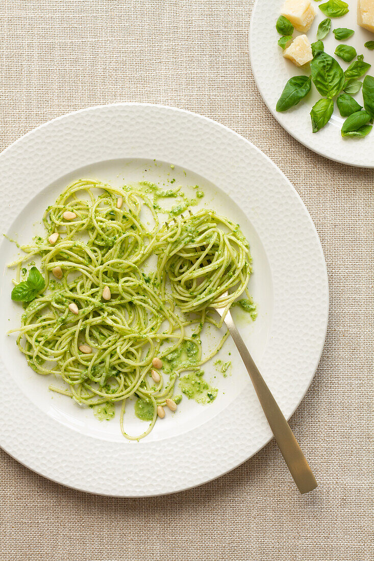 A plate of spaghetti coated in vibrant green pesto sauce, garnished with pine nuts, beside a cluster of basil leaves.