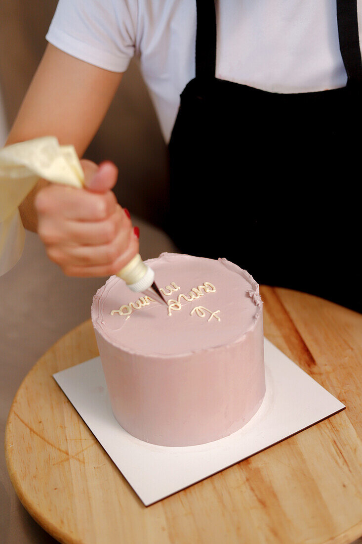 A baker decorates a pink cake with white icing, writing a message in cursive script for a special occasion