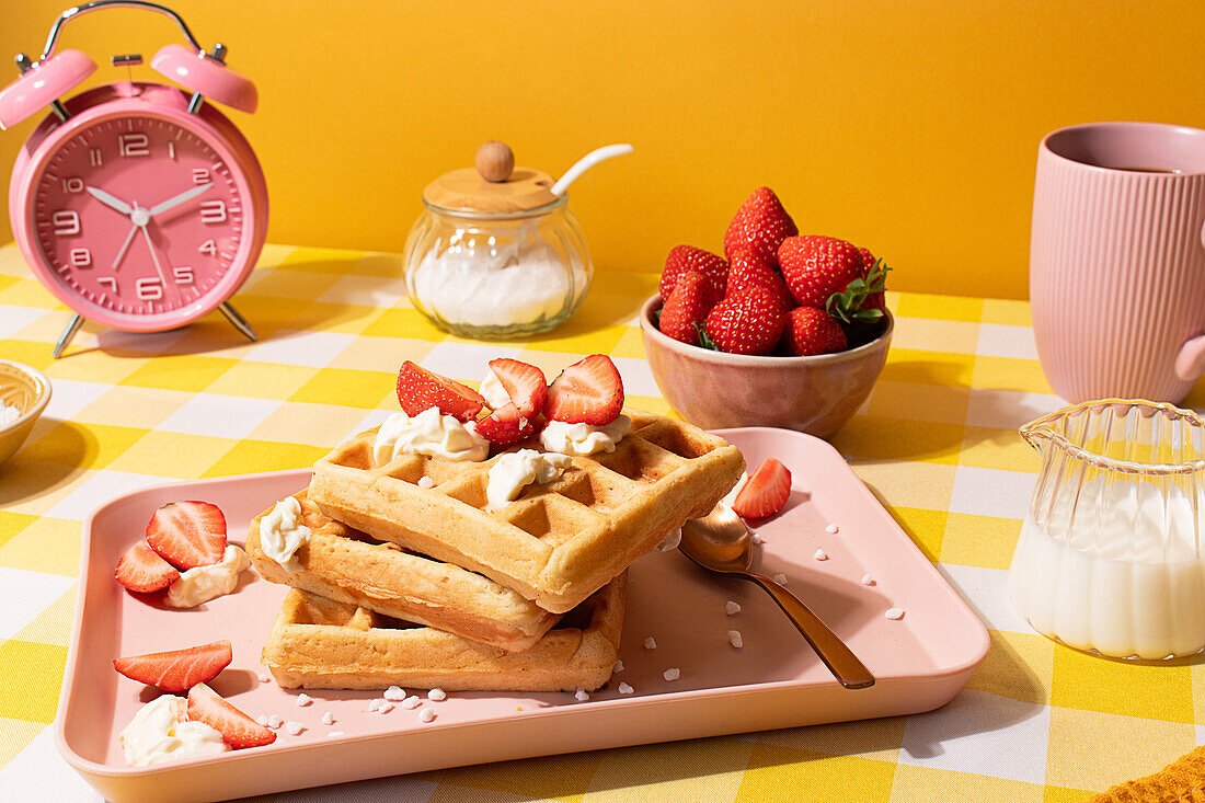 A vibrant still life of waffles topped with whipped cream and fresh strawberries, served on a pink tray against a yellow background, alongside a pink cup and alarm clock