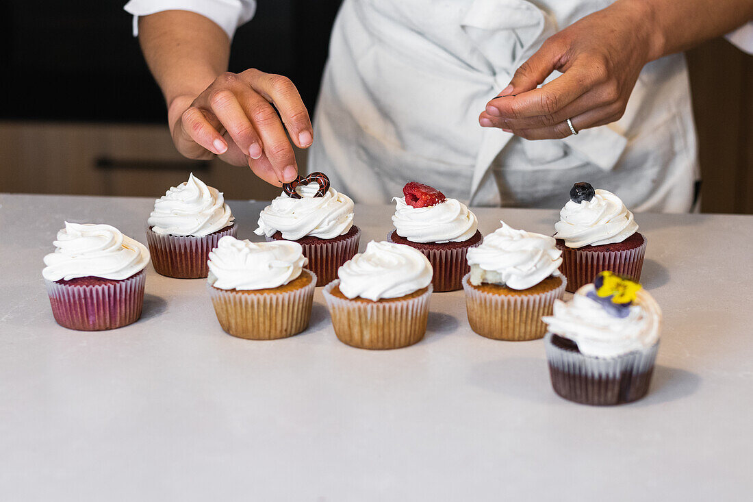 Abgeschnittene, nicht erkennbare Bäckerin, die am Tresen in der Küche einer Bäckerei steht und süße vegane Muffins mit Beeren dekoriert