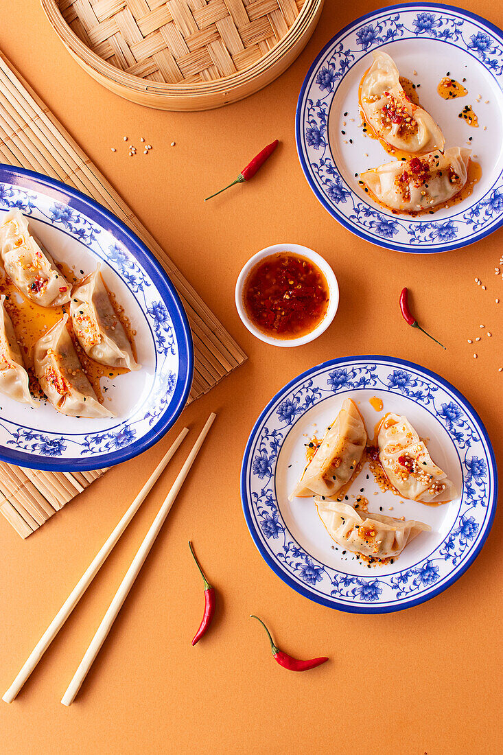 A top-down view of a steamed dumplings garnished with chili flakes and served with a spicy chili dipping sauce, elegantly presented on blue and white decorative plates, accompanied by a bamboo steamer, chopsticks, and a bowl of chili sauce, all set against an orange background