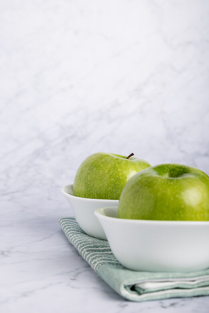 Vibrant green apples placed in elegant white bowls, resting on a soft green napkin set against a clean marble backdrop. This composition highlights wholesome and healthy eating.