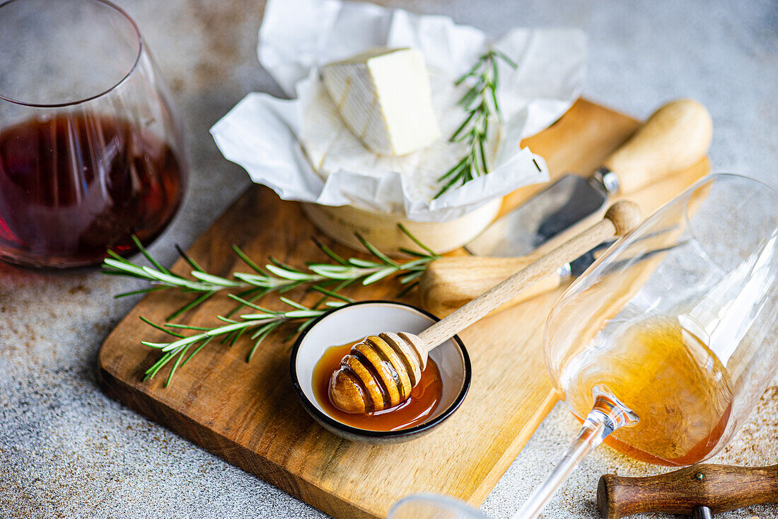 A gourmet selection featuring Brie cheese, fresh rosemary, honey dipper, and glasses of red and white wine, elegantly displayed on a textured backdrop.