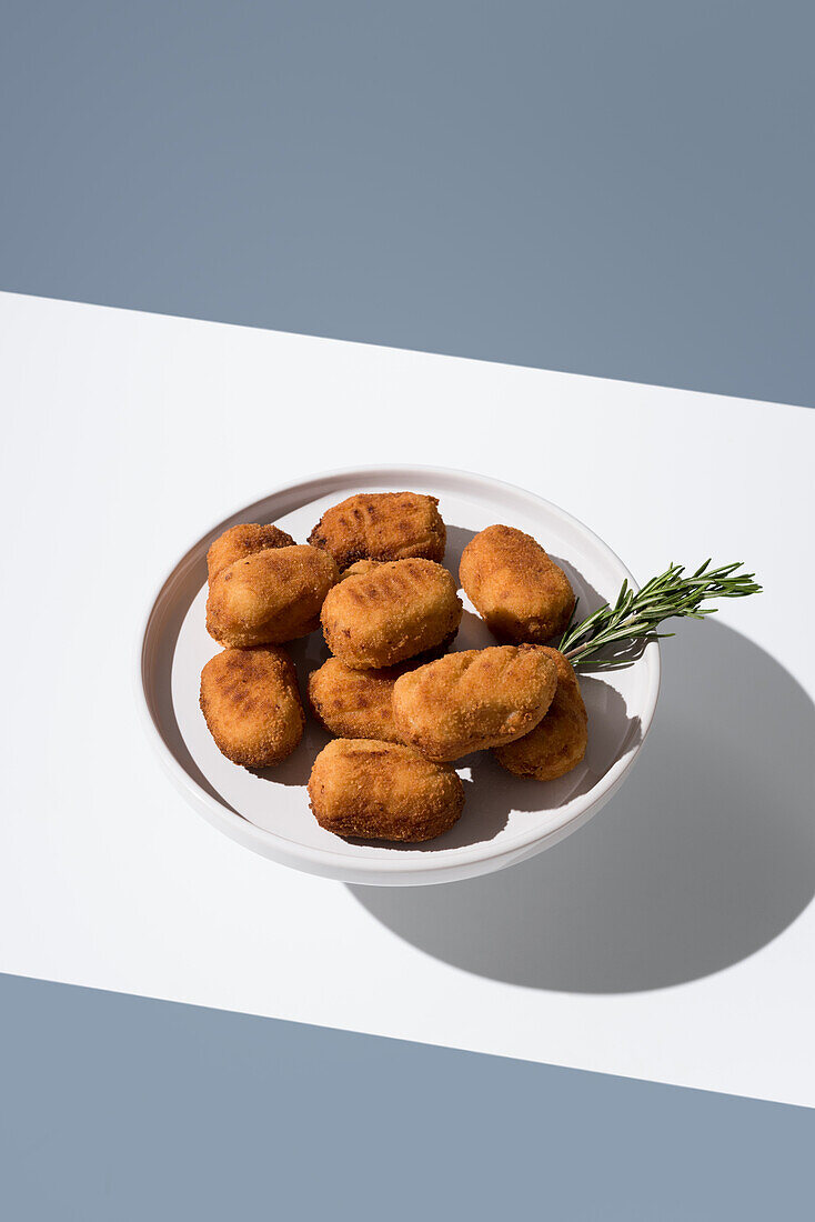 A plate of golden fried croquettes accompanied by a fresh sprig of rosemary, presented on a clean white surface with shadows