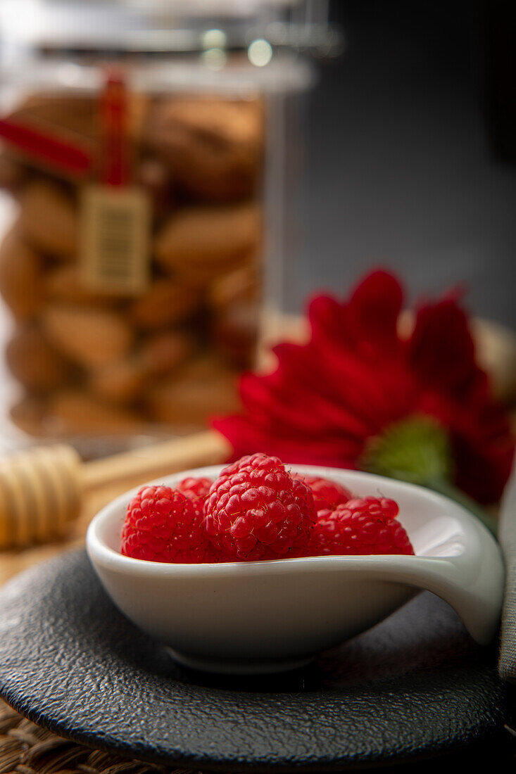 Eine Nahaufnahme saftiger frischer Himbeeren, serviert in einer schlichten weißen Schale, vor einem Weichzeichner-Hintergrund mit einem Behälter mit Keksen und einer leuchtend roten Blume