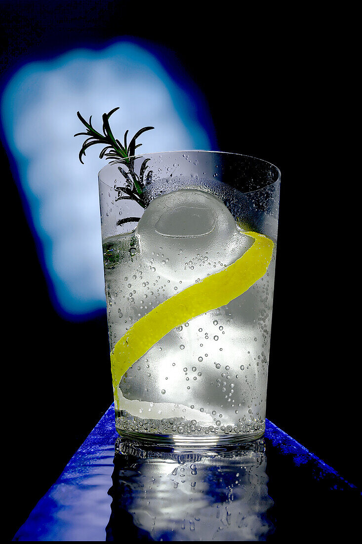 A refreshing gin tonic drink with ice, a twist of lemon peel and a sprig of rosemary, perfectly captured against a blue backdrop