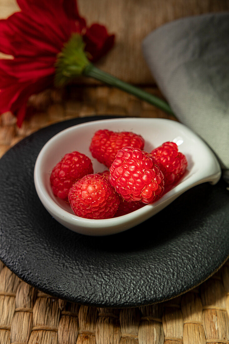 Juicy fresh raspberries artfully arranged in a small white bowl, set against a textured backdrop with a subtle floral element, offering a visually appealing and appetizing scene