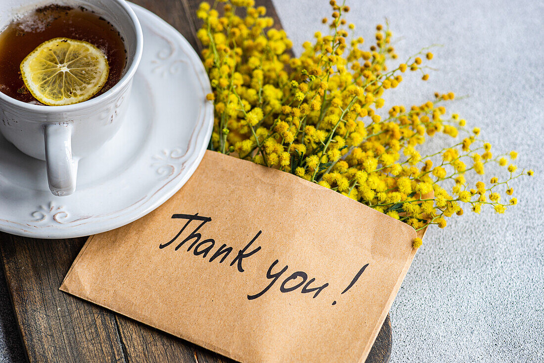 Draufsicht auf eine zarte Tasse Tee mit einer Zitronenscheibe neben einem fröhlichen Strauß gelber Mimosenblüten und einer herzlichen Dankeskarte