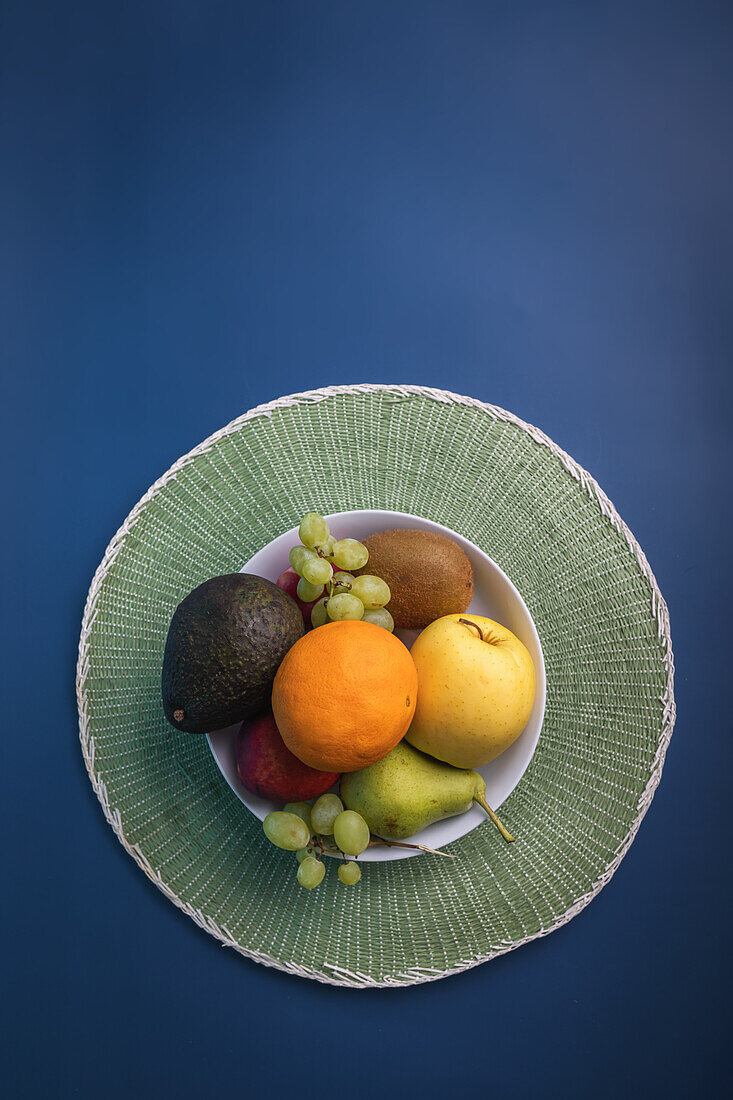 Top view of a colorful array of fresh fruits arranged on a round, textured green placemat against a deep blue background. The composition includes ripe avocado, kiwi, orange, apple, pear, and grapes.