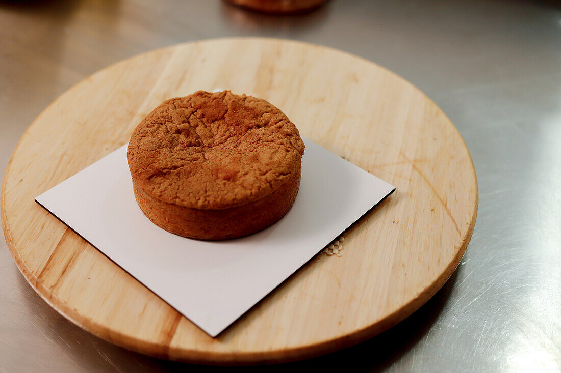 Ein einzelner, frisch gebackener Kuchen, der auf einem hölzernen Schneidebrett abkühlt, vor einer professionellen Küchenkulisse