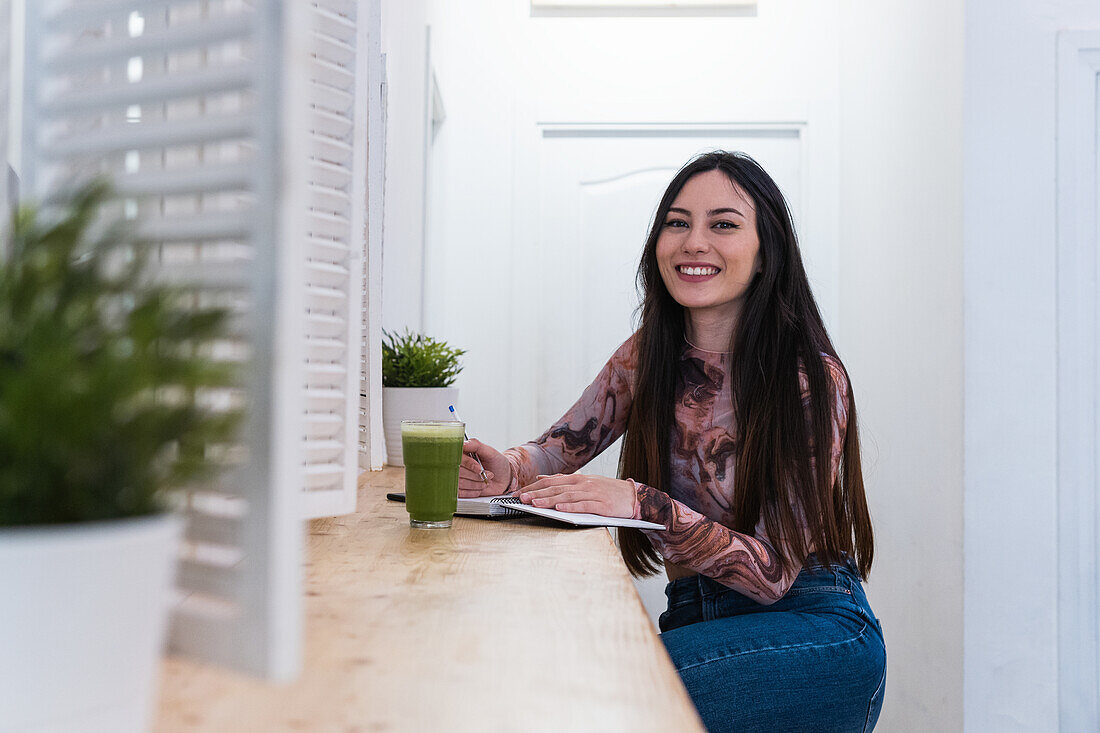 Smiling female sitting at counter with cocktail in bar and writing in notepad while looking at camera