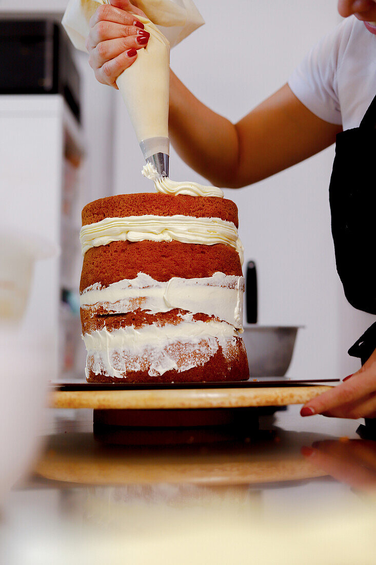 A person applies cream frosting on a multi-layered sponge cake with a piping bag in a kitchen setting