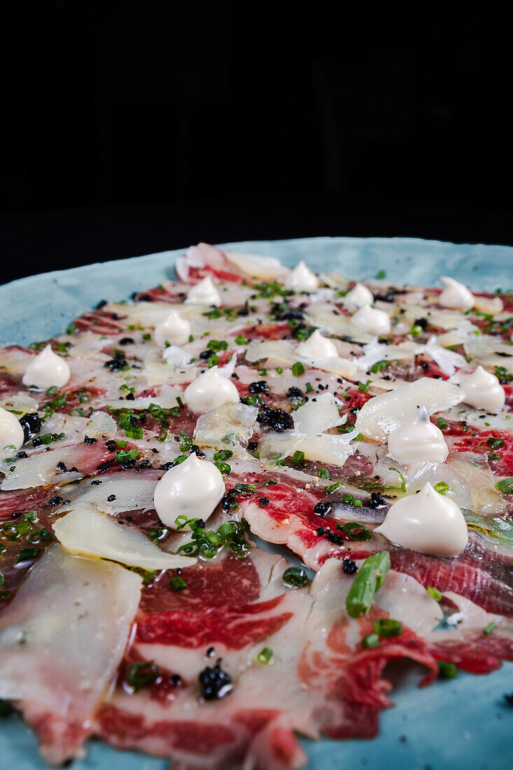 Ein feines Gericht mit Carpaccio aus Serrano-Schinken, garniert mit gehobeltem Parmesankäse, frischen Kräutern und einem Klecks Sahne, elegant präsentiert auf einem blauen, strukturierten Teller.