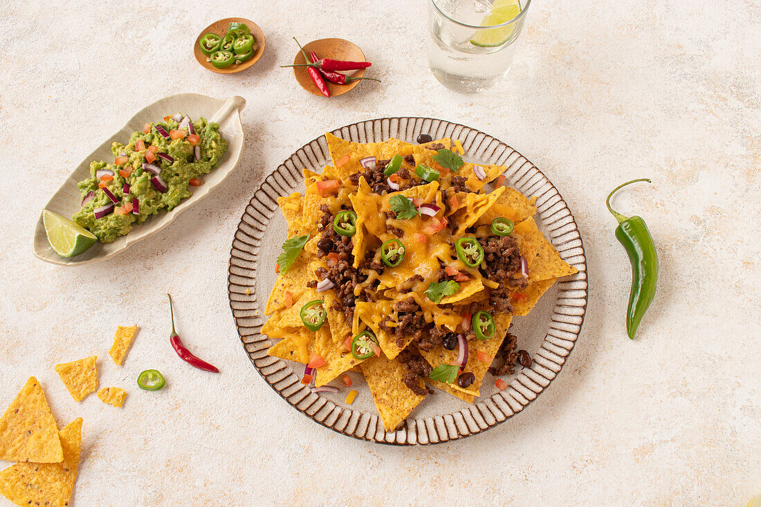 Top view of a platter of nachos generously topped with cheese, ground beef, sliced jalapenos, and fresh tomato, served alongside guacamole and a lime wedge, perfect for snacking or parties.