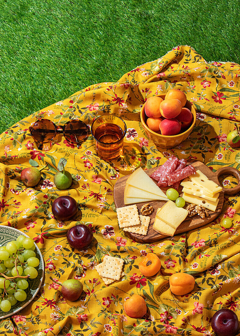 Eine Reihe von bunten Sommerfrüchten und eine Auswahl an Käse und Fleisch, ordentlich präsentiert auf einer gelben, blumengemusterten Picknickdecke, die auf frischem grünen Gras ausgebreitet ist