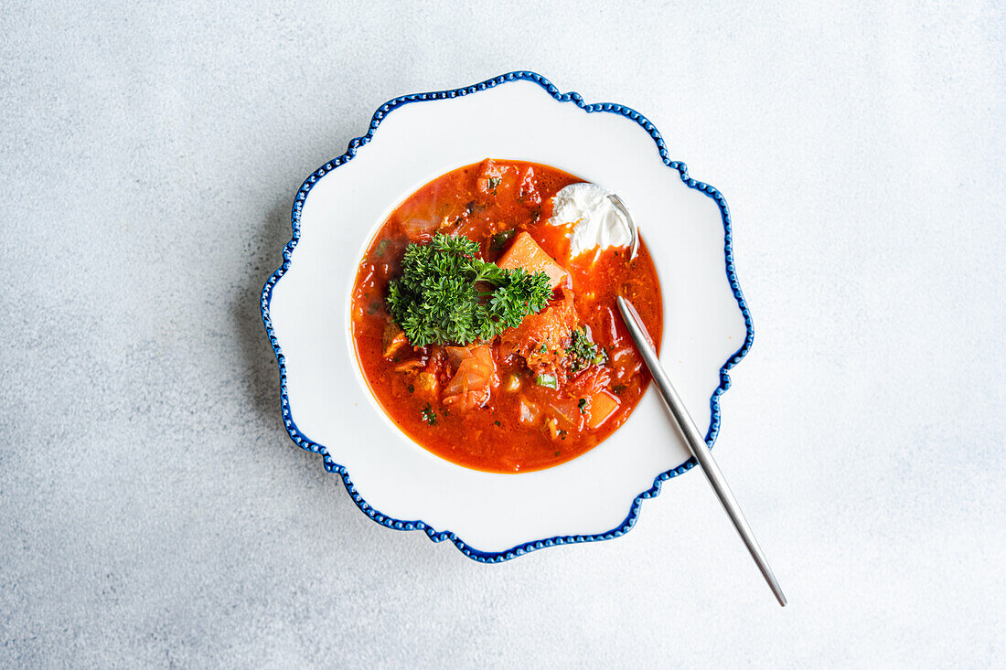 A vibrant bowl of traditional Ukrainian beetroot soup, borscht, with chunks of meat, topped with a dollop of sour cream and fresh herbs, served on a vintage blue-rimmed plate