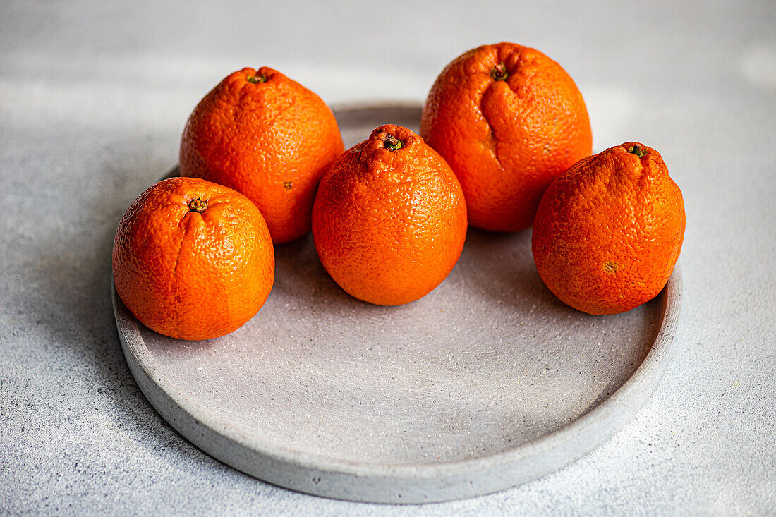 A close-up view of ripe clementines arranged neatly on a round concrete plate, set against a grey background