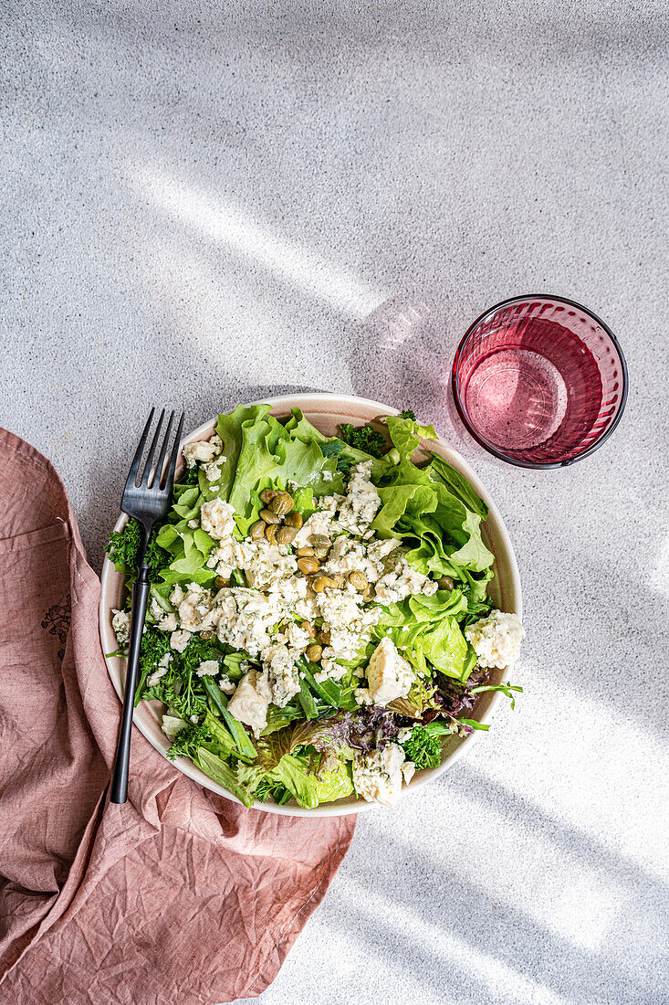 A nutritious herb mix salad containing lettuce, green onion, parsley, tarragon, fermented capers, and crumbled blue brie cheese, drizzled with olive oil and accompanied by a glass of water