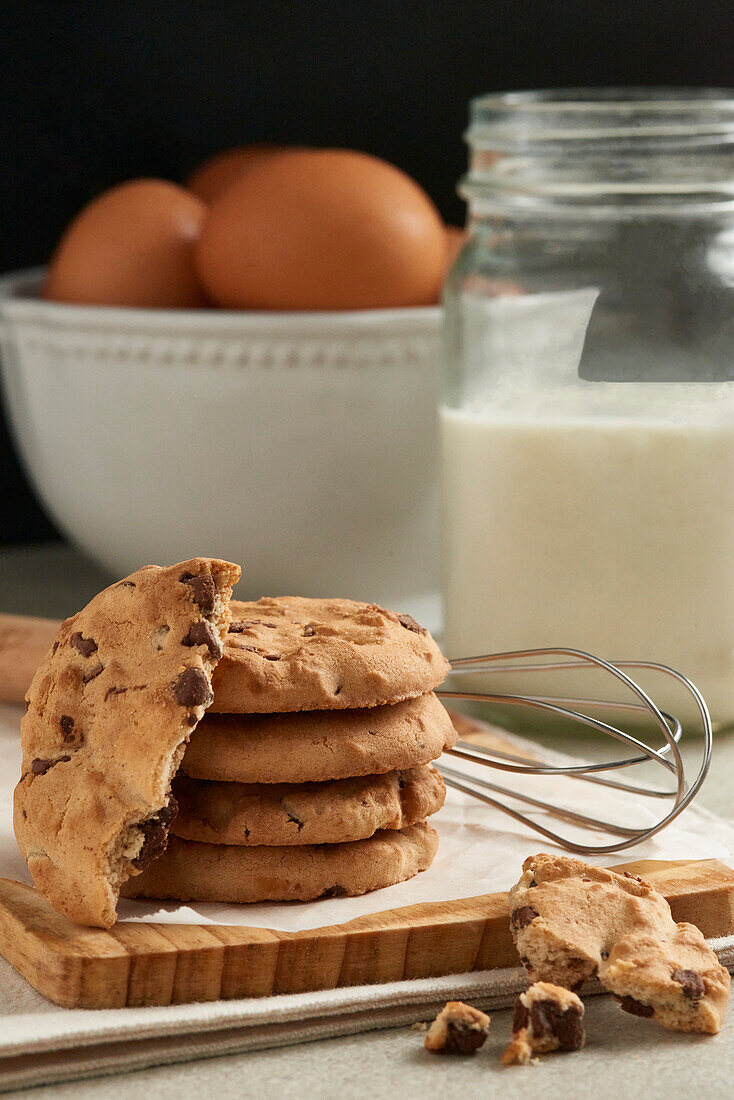 A stack of chocolate chip cookies on a wooden board, with eggs and a jar of milk in the soft-focus background, ready for baking