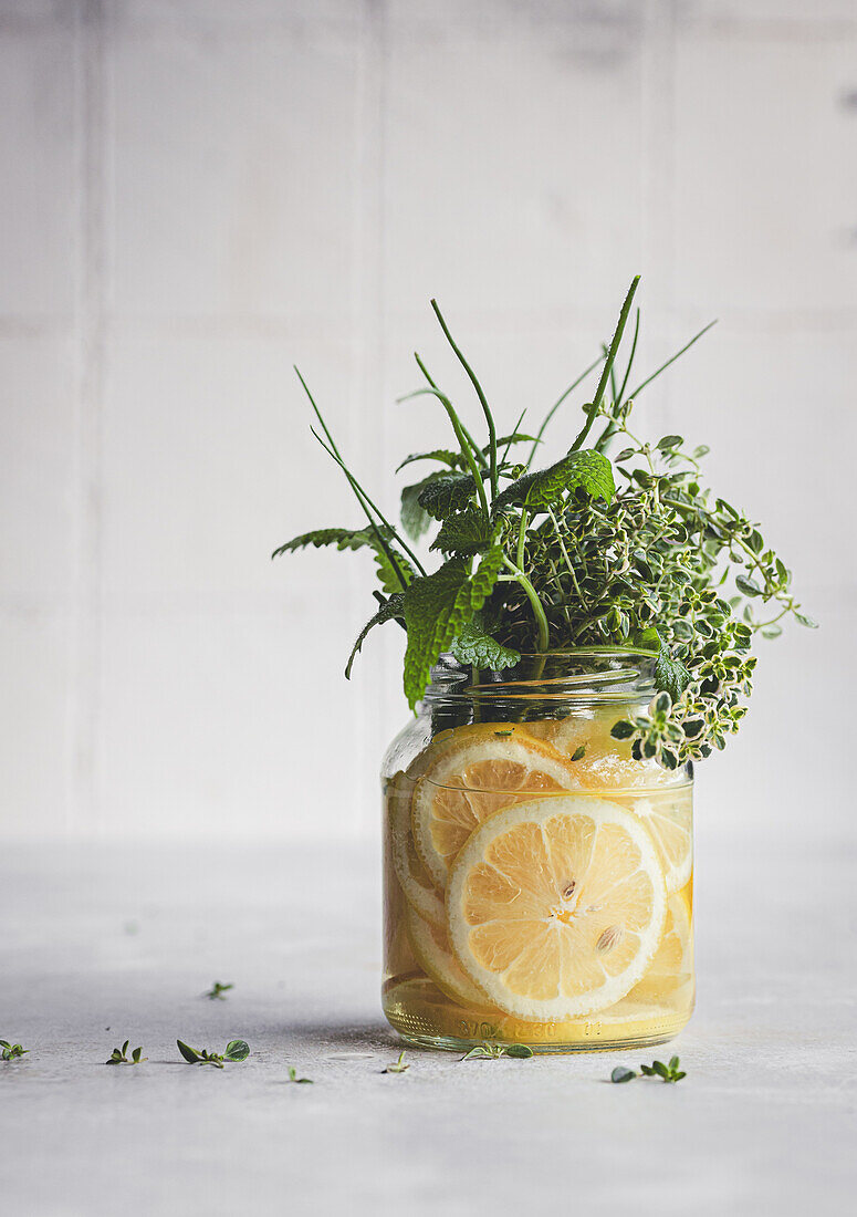 A refreshing jar of infused water filled with sliced lemons and a variety of fresh herbs including lemon thyme, chives, and melissa, set against a neutral background.