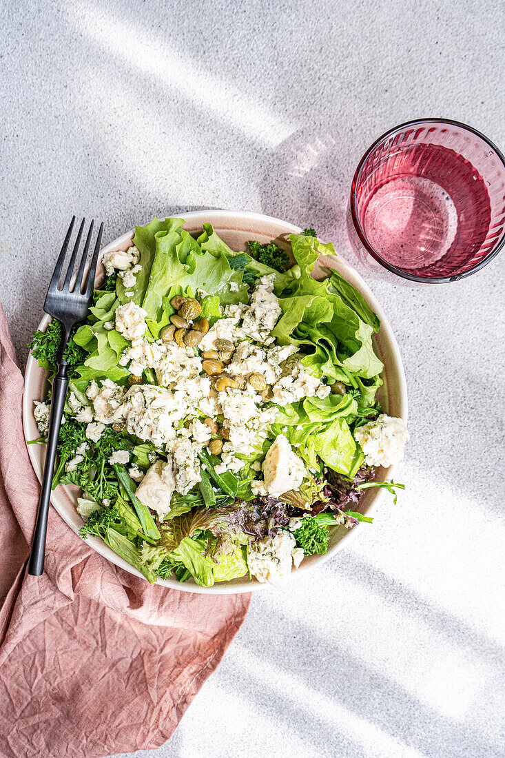 Eine nahrhafte Schüssel mit gemischtem Grünzeug, darunter Kopfsalat, grüne Zwiebeln, Petersilie und Estragon, garniert mit zerbröckeltem Blauschimmelkäse, fermentierten Kapern und einem Spritzer Olivenöl neben einem Glas Wasser