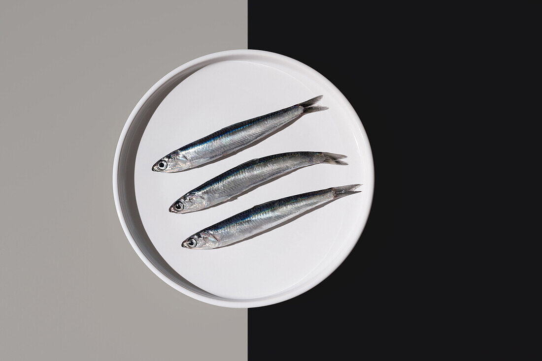 From above of three silvery fresh anchovies aligned on a white round plate, artfully divided by a yin yang background of black and gray