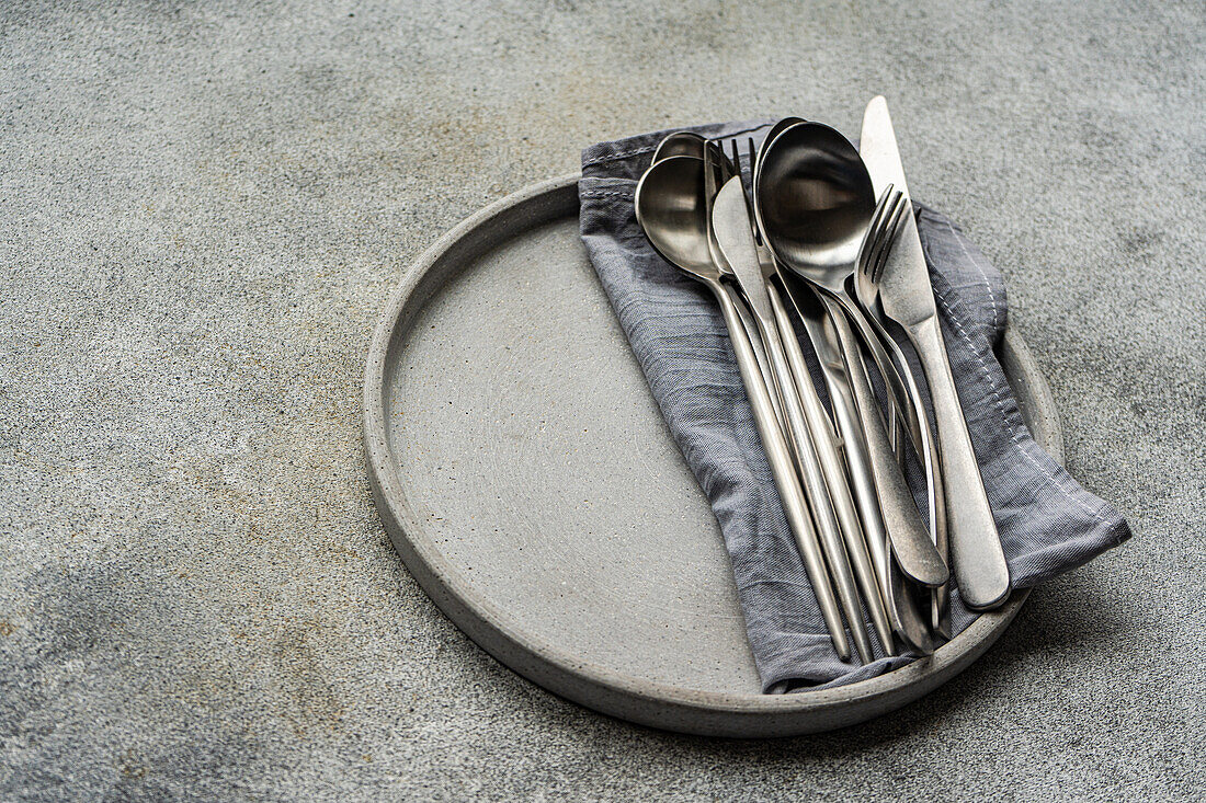 A modern minimalist tableware set up with a ceramic plate, neatly folded grey napkin, and assorted silver cutlery on a concrete backdrop