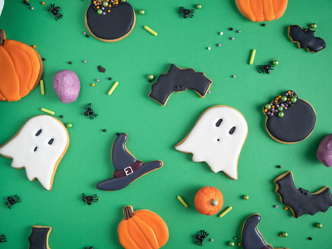 A variety of homemade Halloween-themed cookies, including shapes of ghosts, pumpkins, bats and witch hats, artistically arranged on a bright green background, surrounded by candy beads and decorative spiders