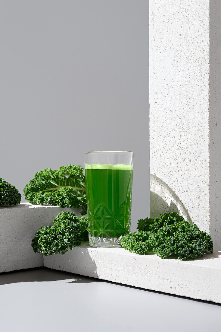 A transparent glass filled with vibrant green kale juice is placed next to fresh, crisp kale leaves on a modern white surface, showcasing a healthful drink option