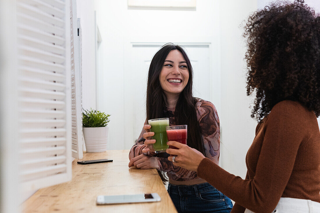 Content multiethnic female friends sitting at counter and drinking refreshing smoothie while chilling in bar and looking at each other