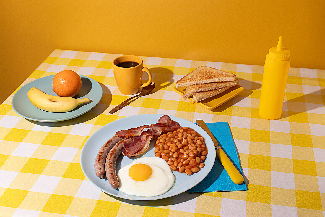 Gelbes Tischtuch mit einem leckeren englischen Frühstück, Ei, Bohnen, Würstchen und Speck, einige getoastete Brotscheiben, Obst und eine Tasse Kaffee