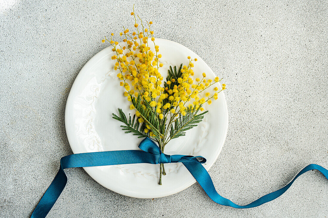 Auf einem verschnörkelten weißen Teller liegt ein leuchtender Strauß Mimosenblüten, elegant gebunden mit einem tiefblauen Band auf einer strukturierten grauen Oberfläche