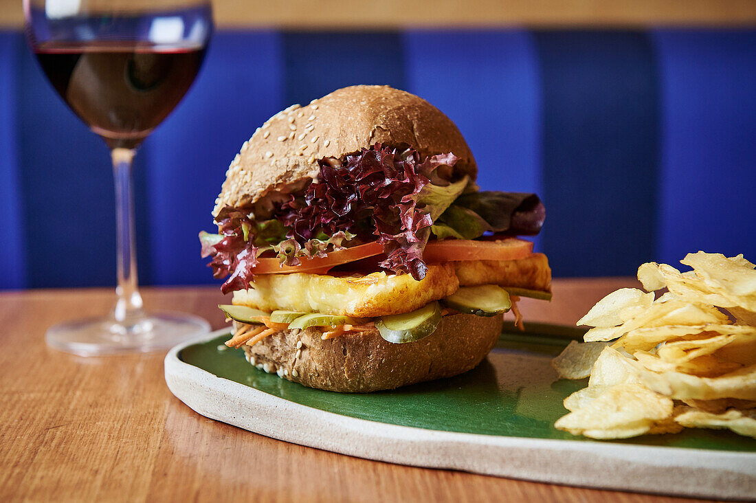 Ein köstlicher Halloumi-Burger, serviert mit knusprigen Pommes frites und einem Glas Rotwein, präsentiert auf einem modernen Keramikteller vor blauem Hintergrund.