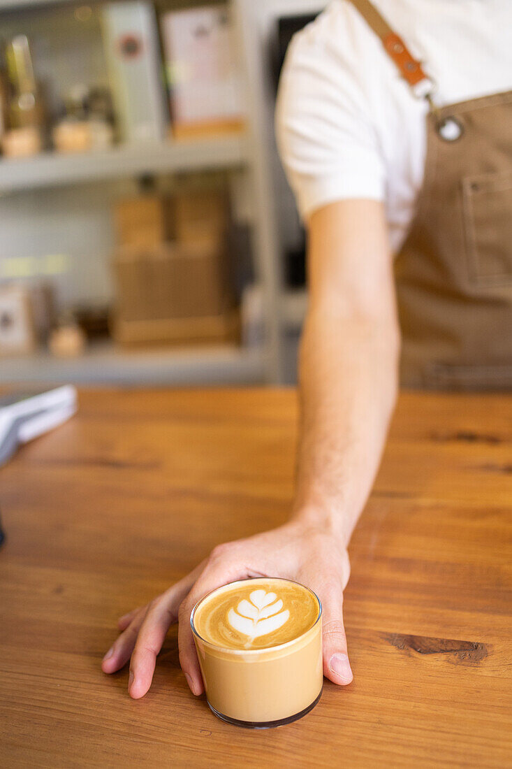 Die Hand eines Barista stellt einen frisch gebrühten Milchkaffee mit Latte Art auf einen Holztresen in einem gemütlichen Café.