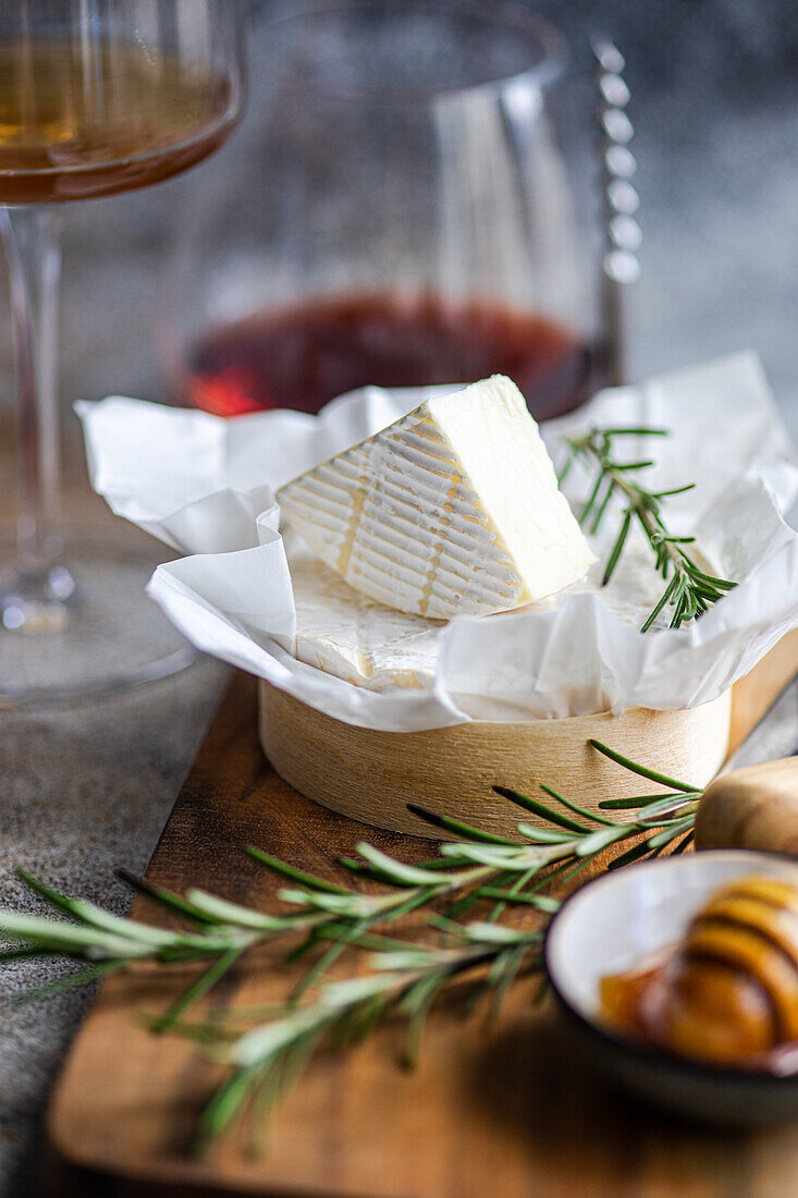 A wedge of Brie cheese on a wooden board, garnished with rosemary, accompanied by wine.