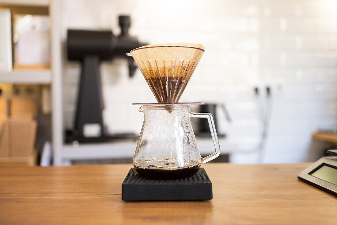 A glass coffee carafe with a drip filter filled with ground coffee, set on a digital scale during a manual brewing process in a modern café environment.