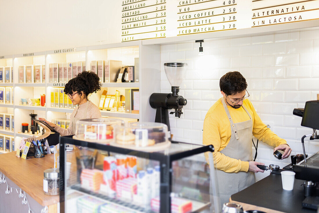 Two multiethnic baristas, one engaged with a tablet and the other preparing coffee, in a bright, contemporary cafe with a visible price list.