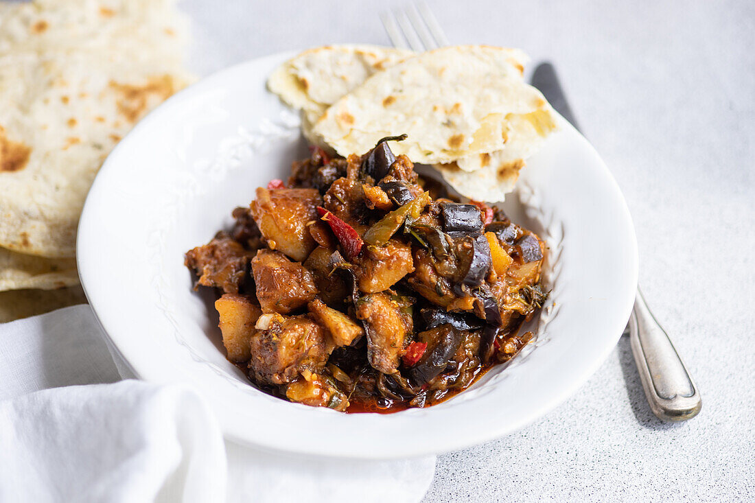 Ein köstlicher Gemüseeintopf mit Auberginen, Tomaten, Kartoffeln, Paprika, Zwiebeln, Knoblauch und Basilikum, begleitet von frisch gebackenem Fladenbrot auf einem weißen Teller