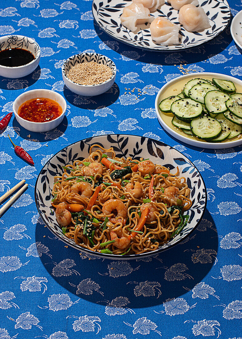 Top view of a lively table set with Chinese cuisine, featuring stir-fried noodles with shrimp and vegetables in a traditional decorative bowl, surrounded by various dips, sesame seed sprinkled dumplings, sliced cucumbers, and red chili peppers