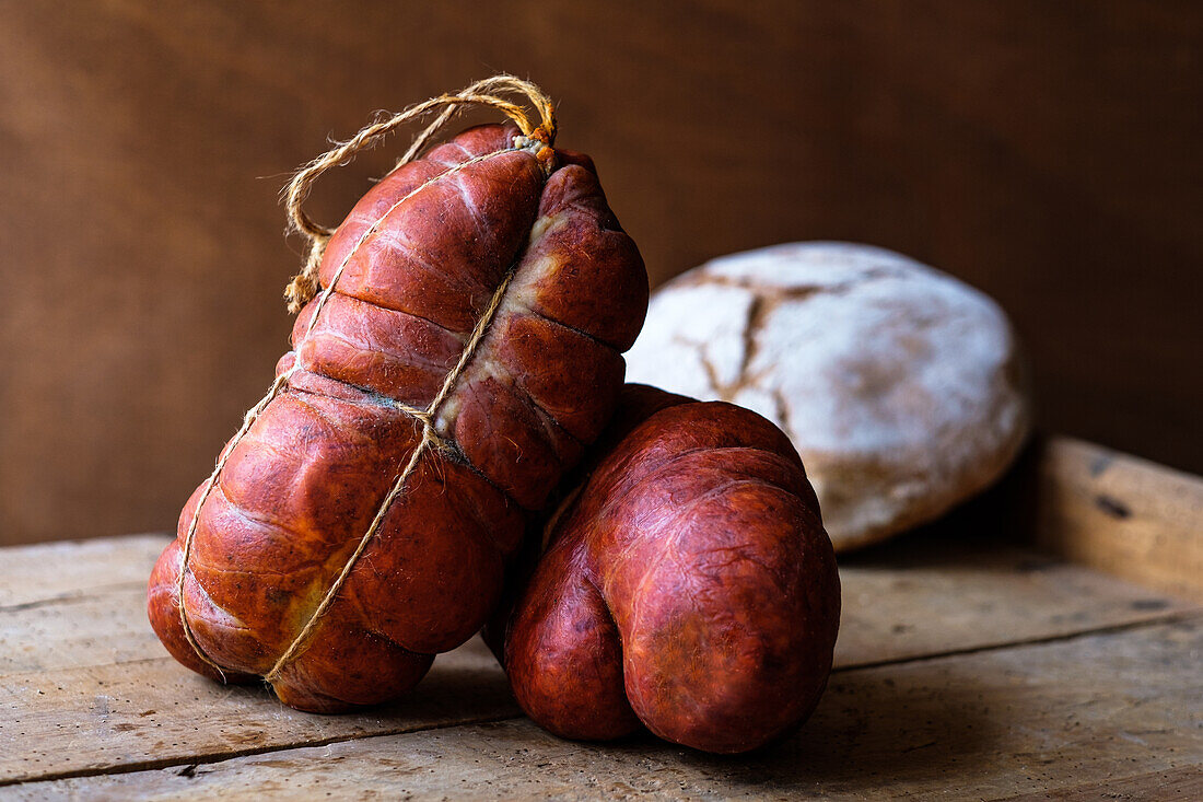 Eine Auswahl traditioneller spanischer Wurstwaren, allen voran die Sobrasada, eine rohe, gepökelte Wurst von den Balearen, die aus gemahlenem Schweinefleisch, Paprika und Gewürzen besteht. Die Würste sind mit Bindfäden zusammengebunden und liegen auf einem rustikalen Holzbrett mit einer anderen Wurstsorte im Hintergrund.