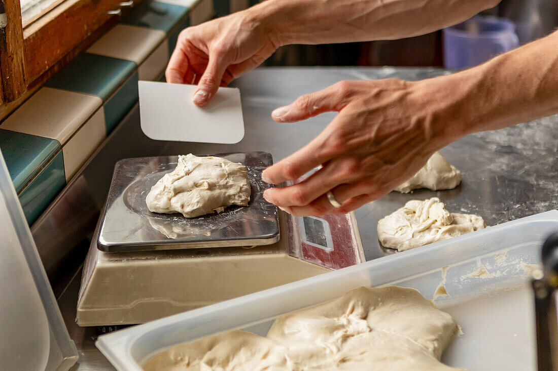 A person measures and prepares fresh pizza dough on a digital scale in a professional kitchen, surrounded by containers of dough and baking tools