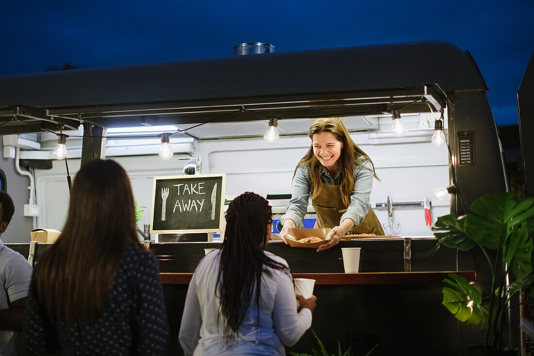 Happy adult woman in apron giving takeaway box with dish to black female client while working in food trunk at night