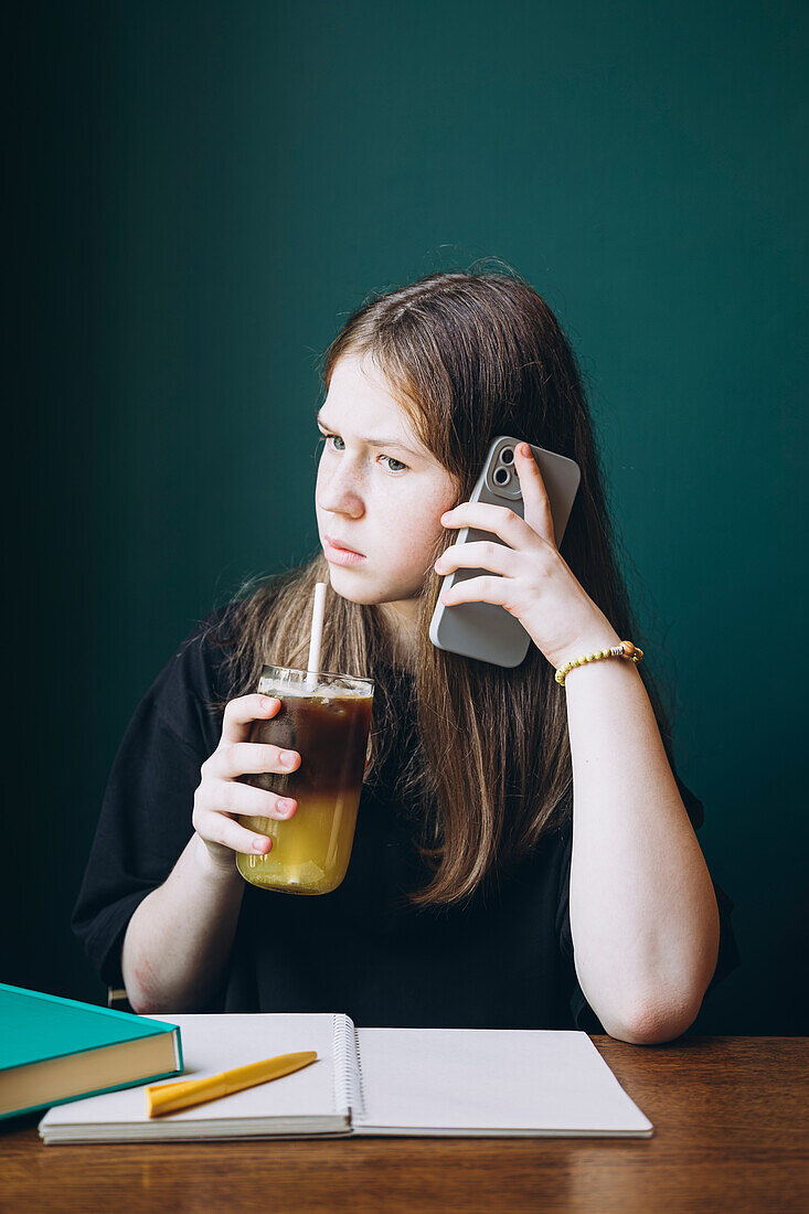 A young female student takes a break from studying to have a drink and talk on her smartphone. She is looking away from the camera, highlighting a moment of distraction or pause in her study routine.