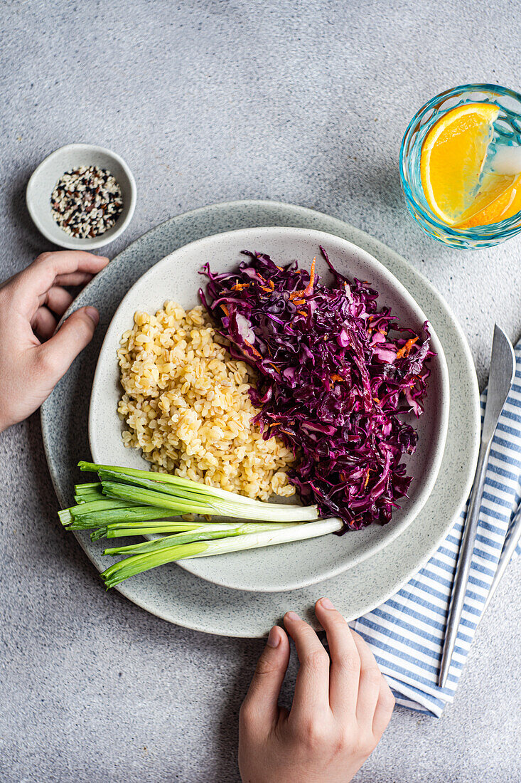 A nutritious lunch featuring a vibrant salad of raw red cabbage and carrot alongside fluffy boiled bulgur, served on a ceramic plate with fresh green onions and a citrus water on the side