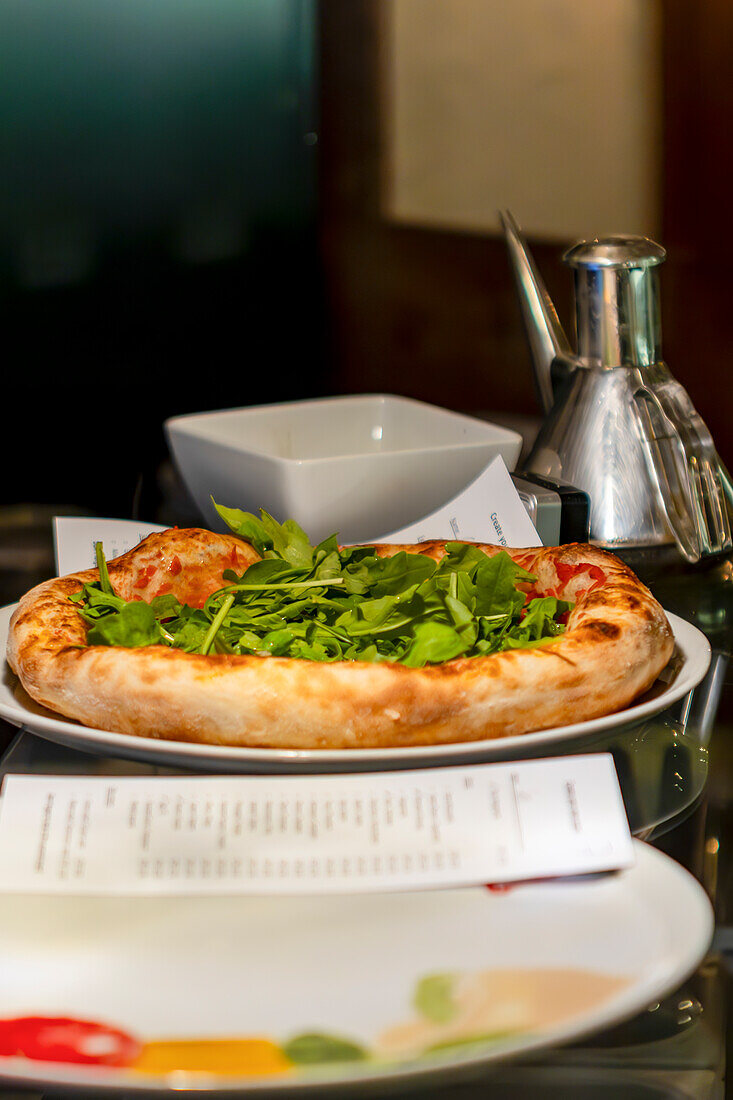 A tantalizing freshly baked pizza, generously topped with fresh arugula, served on a white plate in a stylish restaurant setting, accompanied by a metallic oil dispenser and a white ceramic bowl