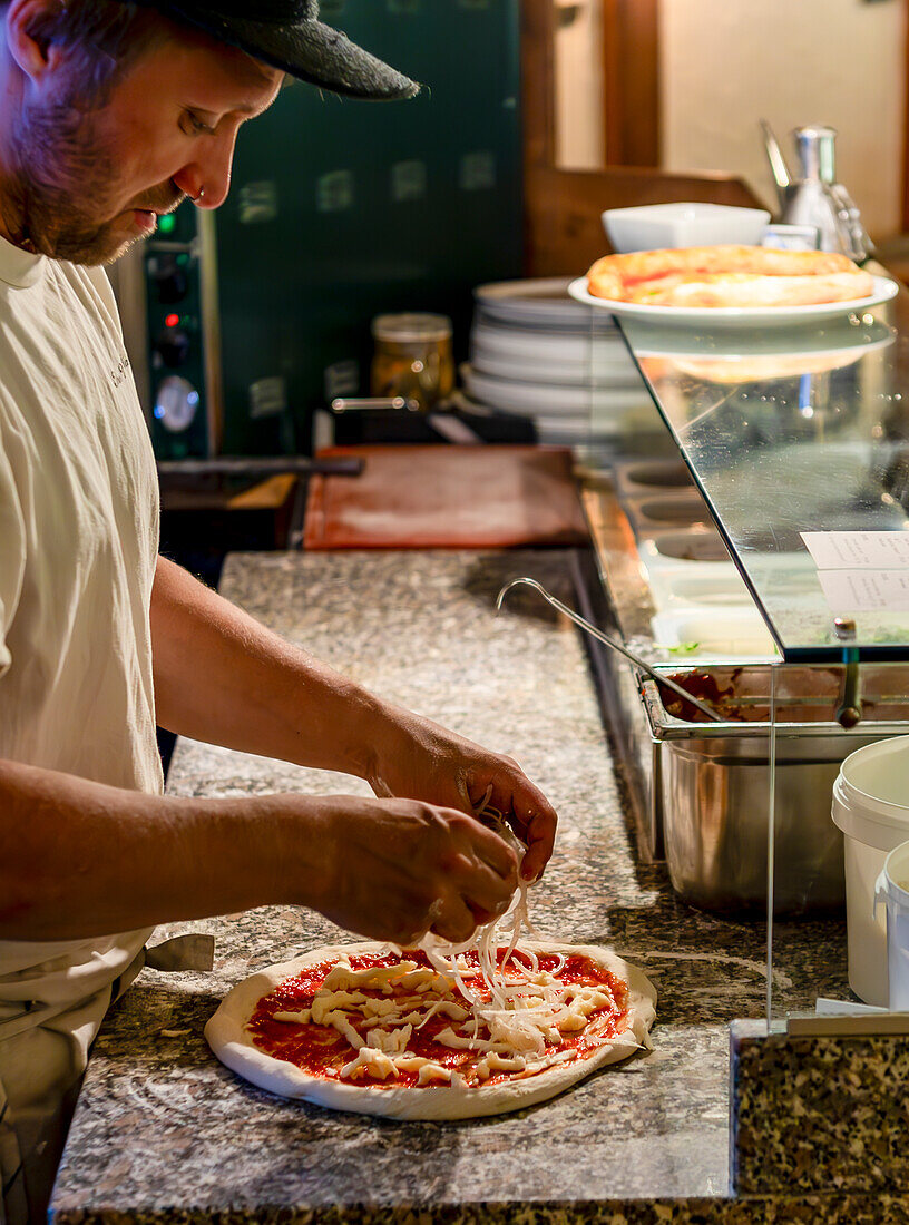 Ein Profikoch gibt in einer gut ausgestatteten Großküche vorsichtig Käse auf einen frisch belegten Pizzaboden und zeigt die Kunst der Pizzaherstellung