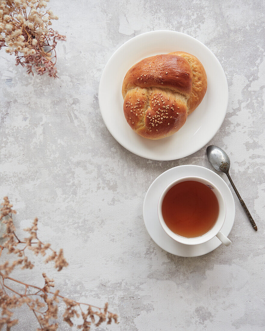 Draufsicht auf ein herbstliches Teeservice mit einer Tasse heißem Tee und einem frisch gebackenen Sesambrot auf einer strukturierten Oberfläche, begleitet von Trockenblumen