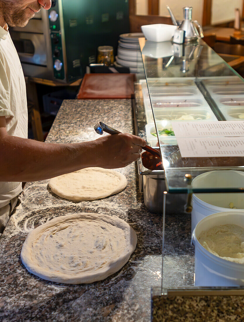 A skilled pizza chef in action, spreading sauce meticulously over fresh pizza dough in a restaurant kitchen, surrounded by various toppings and tools