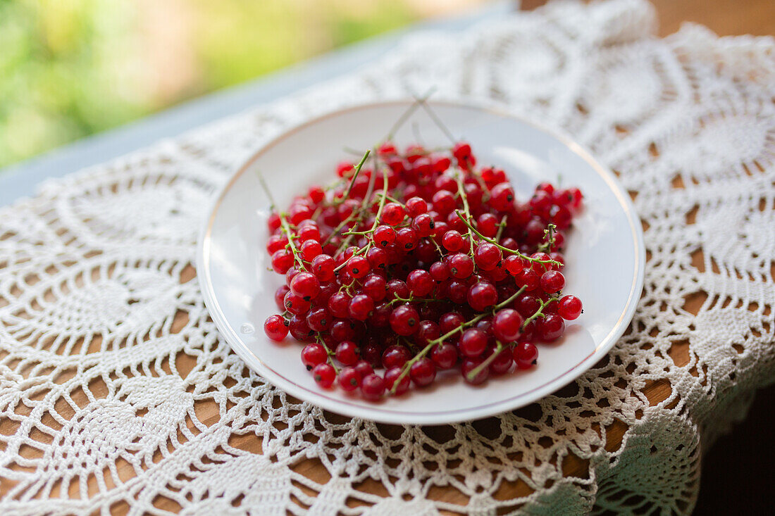 A bowl of ripe redcurrant berries on a beautifully patterned white lace tablecloth, sitting beside a window with natural light